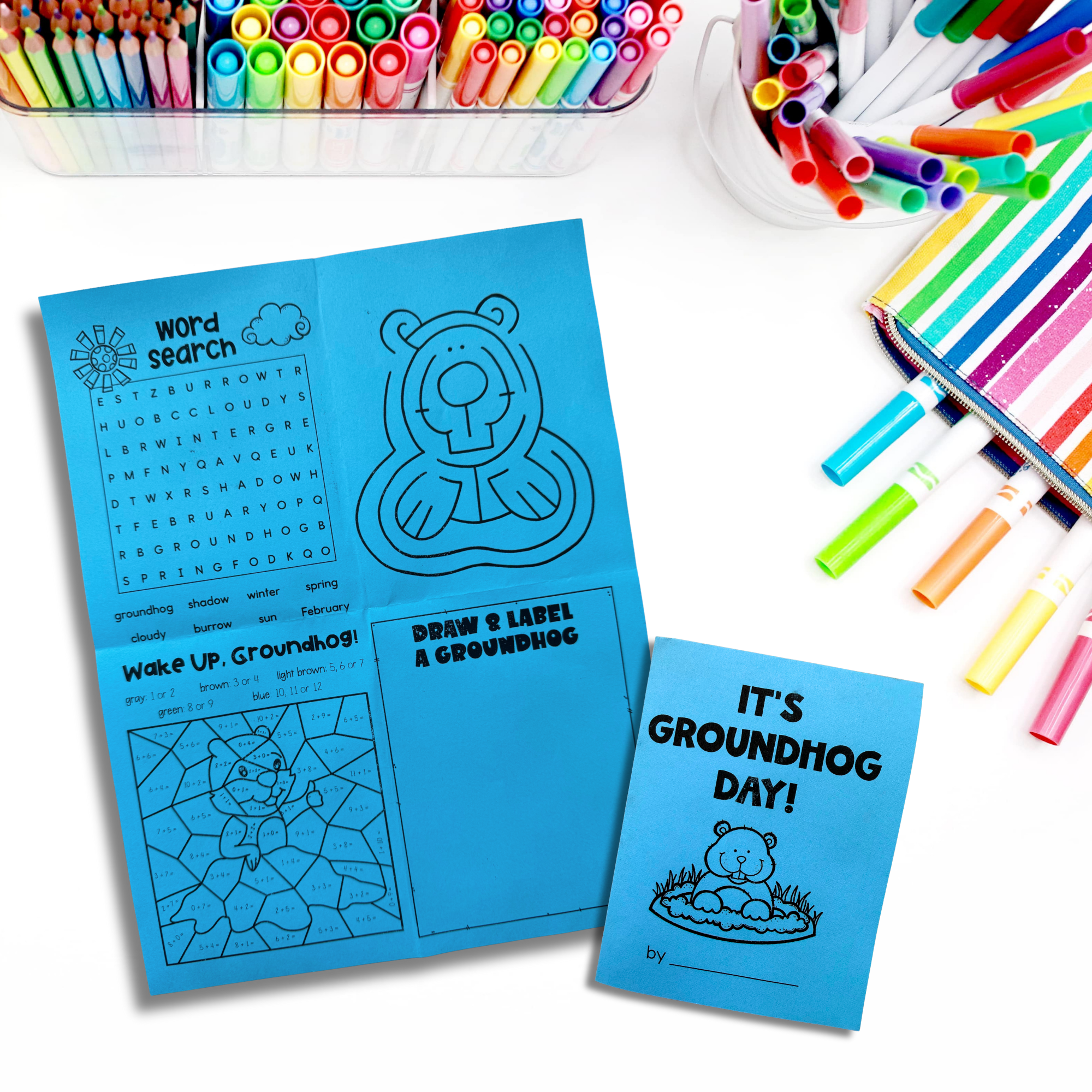 A blue worksheet and booklet about Groundhog Day with activities, markers, and colored pencils arranged on a white surface.