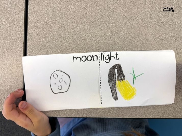 A child's drawing explores compound words, displaying "moonlight" above two images: a moon on the left and a yellow light with a black object on the right. A child's hand holds the paper.