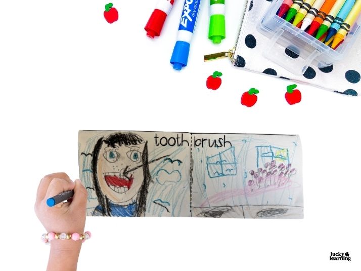 A child’s hand draws in a booklet labeled “tooth: brush,” exploring compound words, with markers, crayons, and apple erasers arranged above on a white surface.