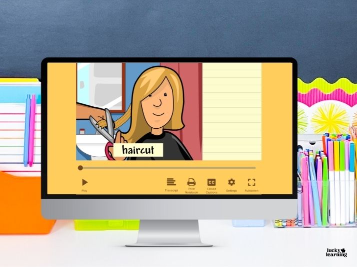 A computer screen displays an animated video of a person getting a haircut, with the compound word "haircut" labeled, on a desk with colorful stationery in the background.