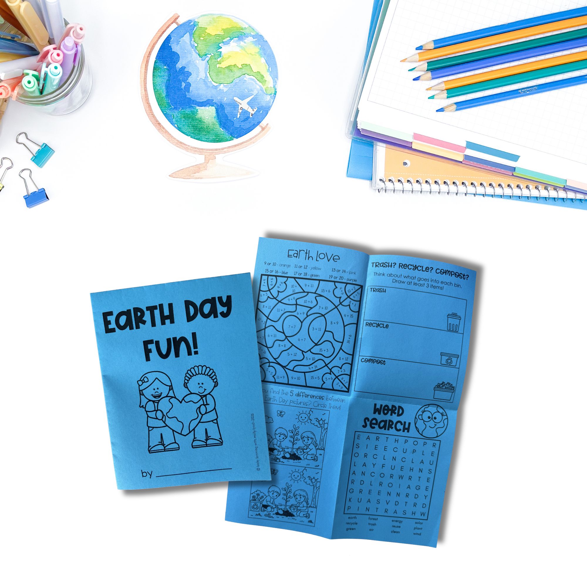 A booklet titled "Earth Day Fun!" is open to activity pages, placed on a white desk with colored pencils, notebooks, clips, and a painted globe.