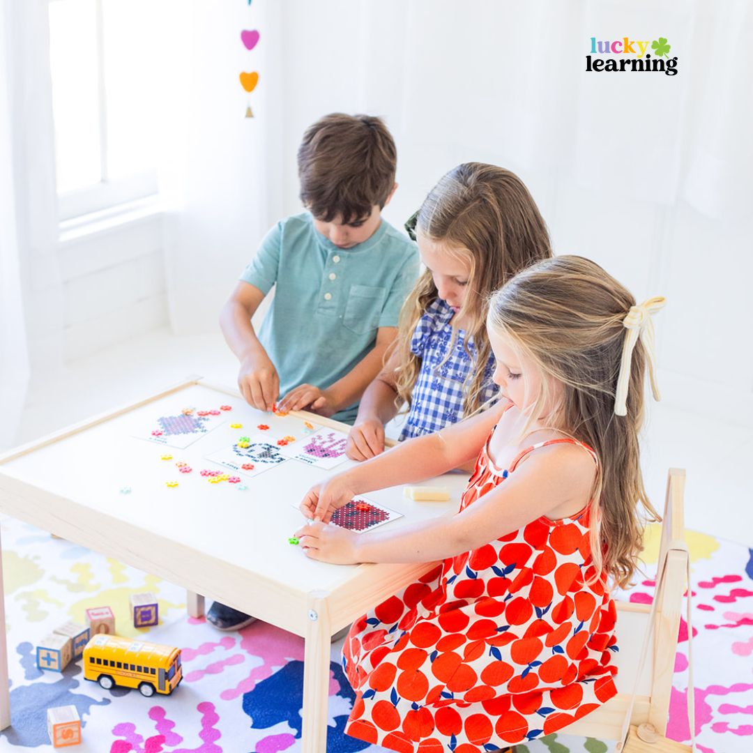 Three young children sit around a small table, focused on arts and crafts activities with colorful beads and shapes, while math fact fluency games add an extra layer of fun. A toy school bus is on the floor nearby.