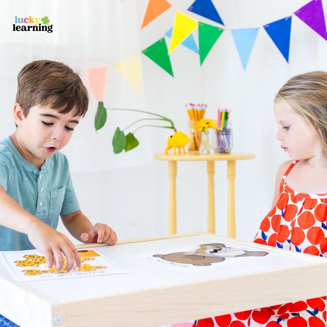Two young children sit at a table, working on educational activity sheets and math fact fluency games. Colorful pennant banners and art supplies are visible in the background.