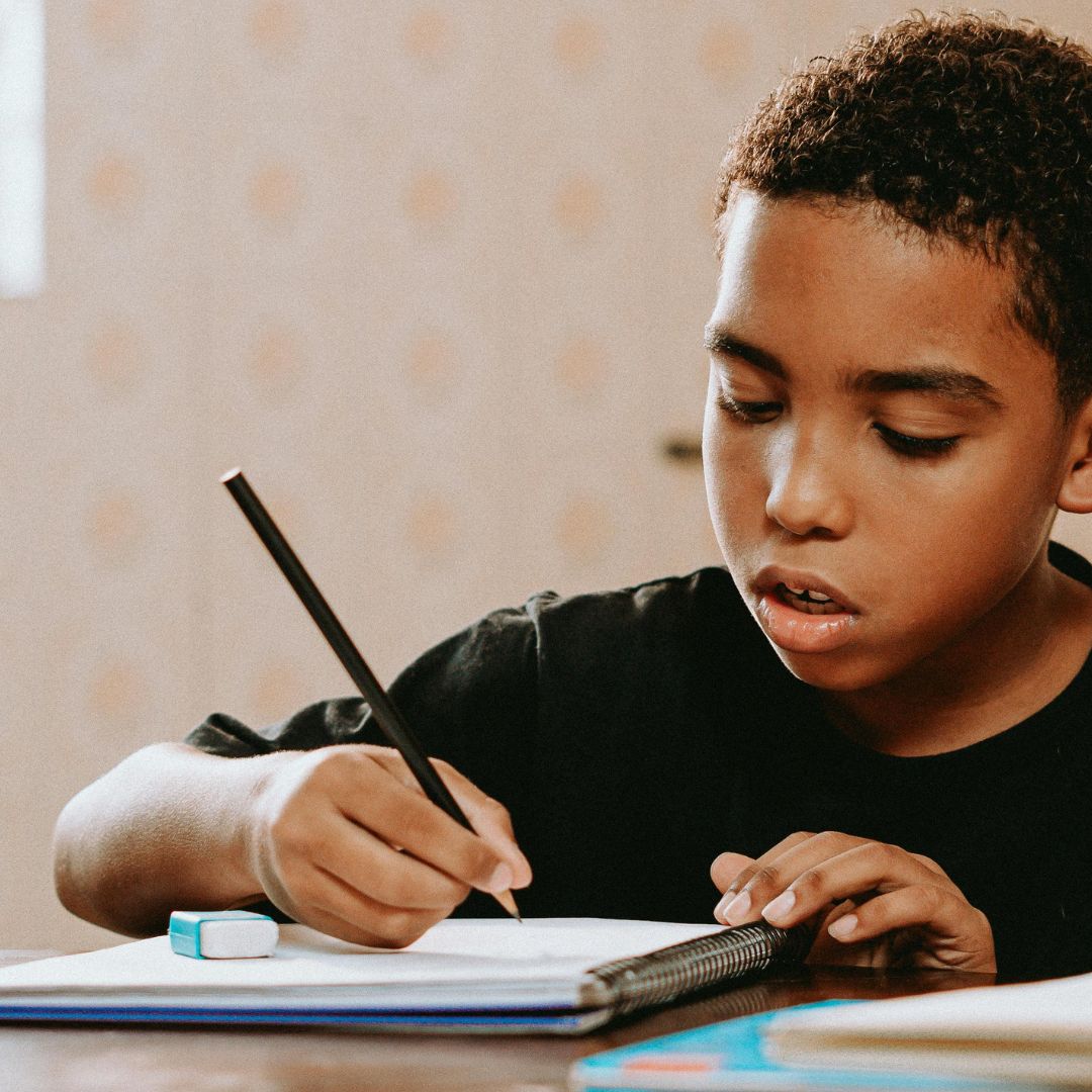 A boy sits at a table writing in a notebook with a pencil, an eraser and another notebook nearby, practicing after playing math fact fluency games.