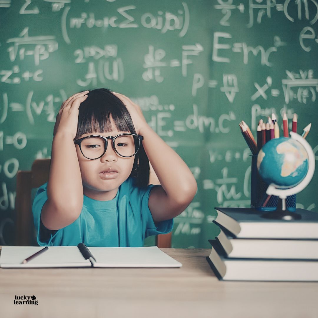 Young child in glasses looks frustrated at a desk with books and a globe, struggling with following directions in front of a chalkboard filled with math equations.