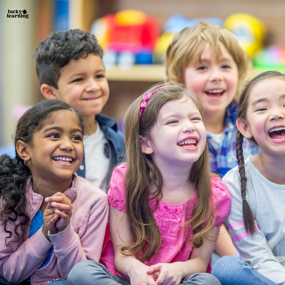 Five young children sit close together indoors, smiling and laughing as they enjoy an activity that involves following directions. The background is blurred, with colorful objects visible.