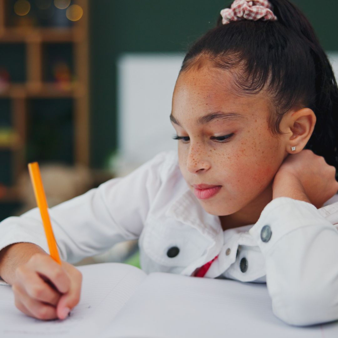 A young girl with a ponytail writes in a notebook with a pencil, resting her head on her hand and wearing a white jacket.