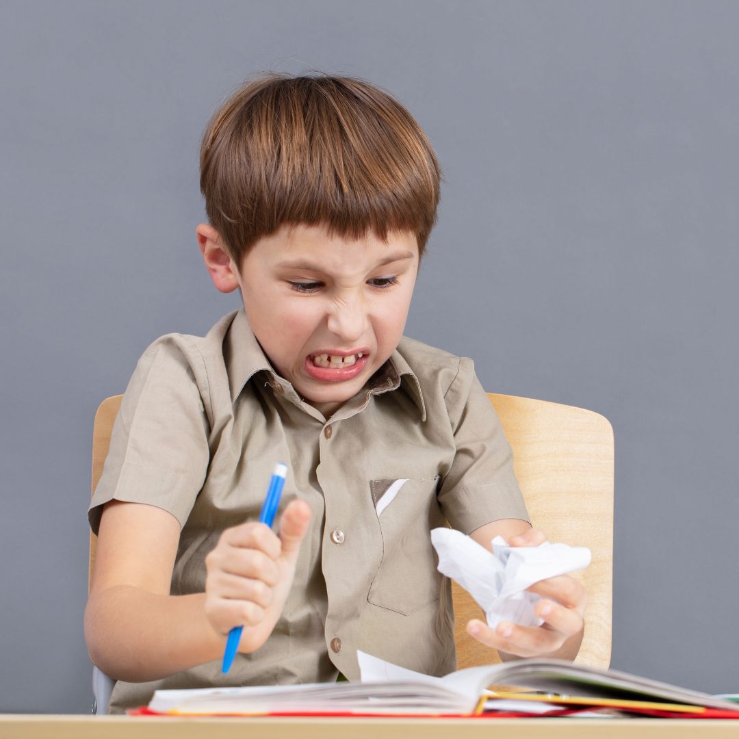 A young boy sitting at a desk clenches a crumpled piece of paper in one hand and a pen in the other, showing a frustrated facial expression.