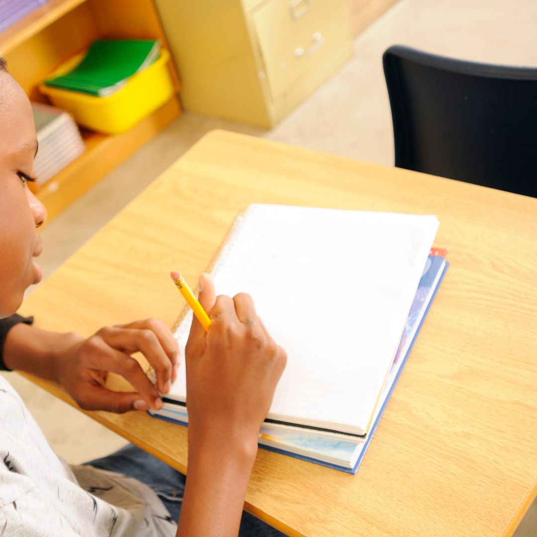 A student sits at a desk holding a pencil and writing in a blank notebook, with books and shelves visible in the background.