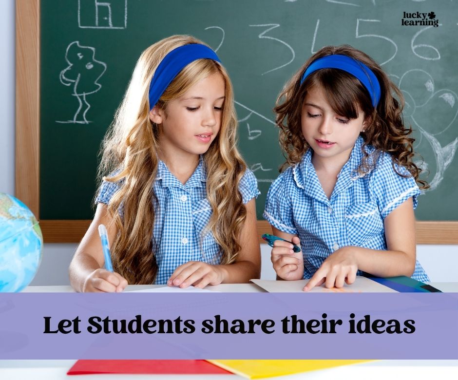 Two young girls in blue uniforms sit at a desk writing and discussing journal writing prompts for first grade in front of a chalkboard, with the text “Let Students share their ideas” displayed below.