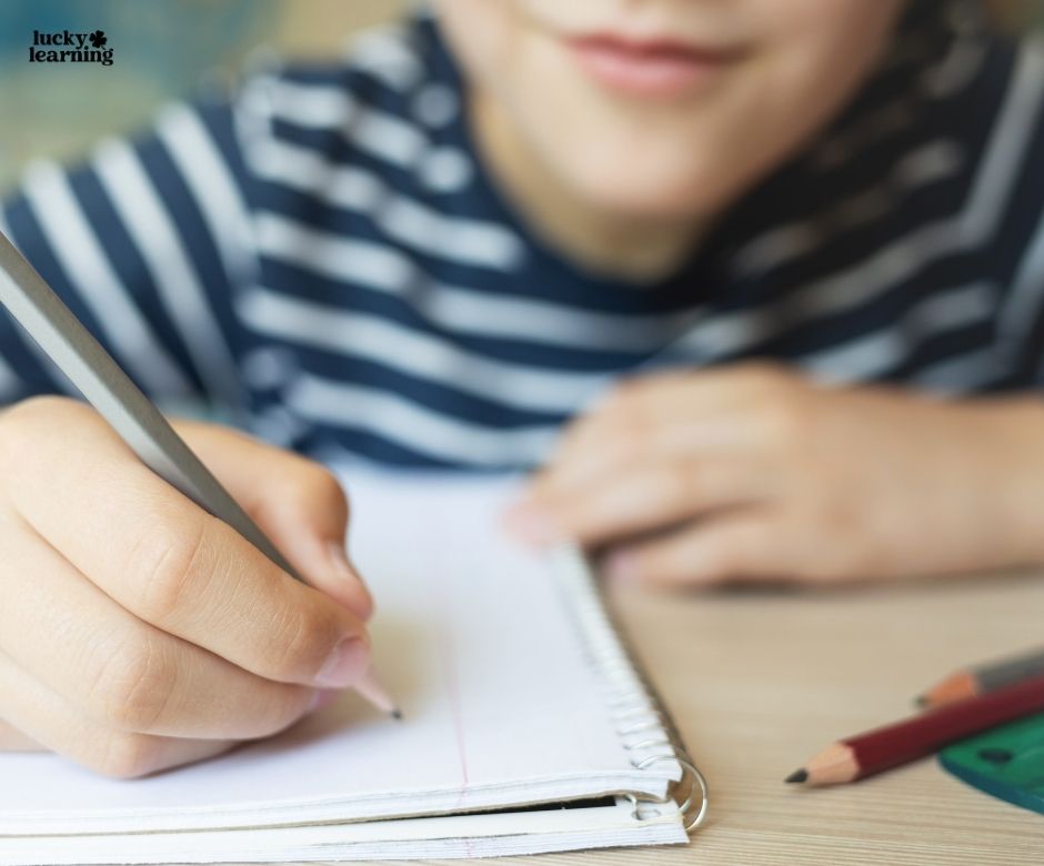 A child wearing a striped shirt writes in a spiral notebook with a pencil, perhaps working on journal writing prompts for first grade; colored pencils are on the desk.