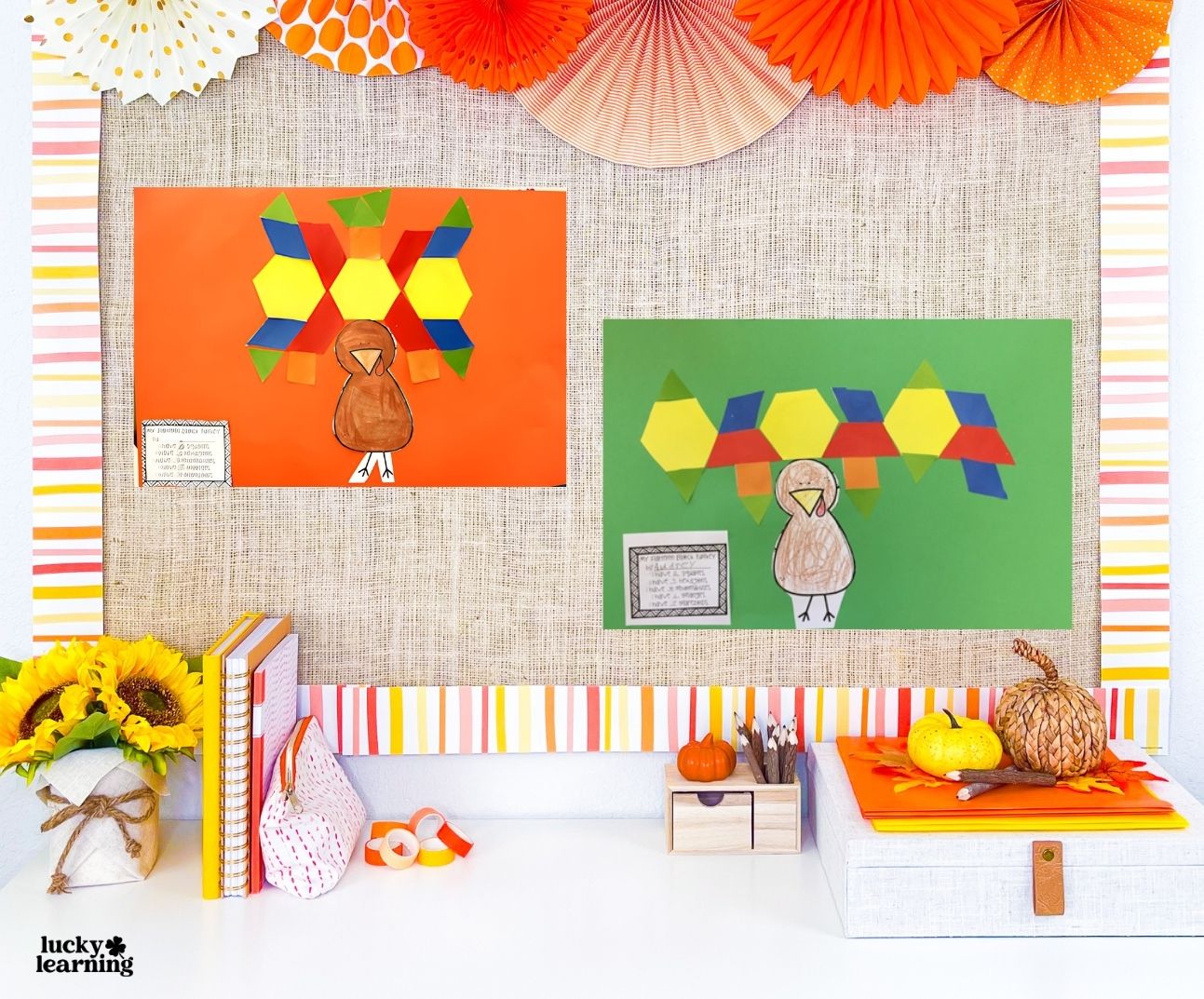 Two colorful turkey crafts made from paper shapes, inspired by pattern block activities, are displayed on a bulletin board, with small pumpkins, books, and autumn decorations arranged on a white desk below.