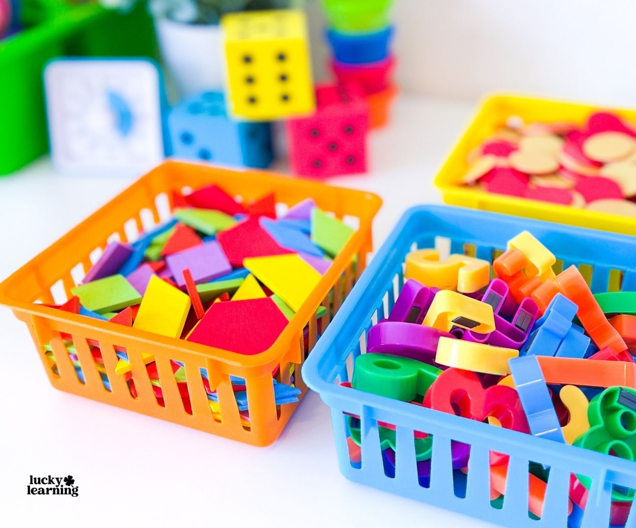 Two plastic baskets filled with colorful geometric shapes and plastic alphabet letters, perfect for engaging pattern block activities, with more math and learning toys visible in the background.