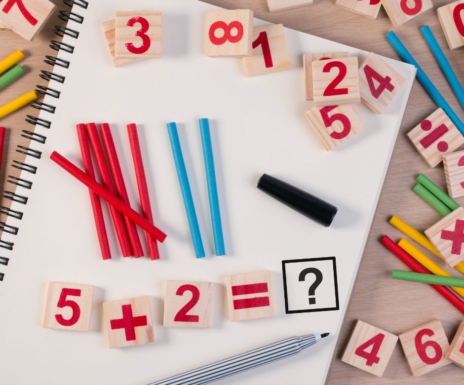 Math puzzle with wooden number blocks, colored sticks, and a pen on a notebook; equation 5 + 2 = ? is displayed with blank space for the answer.