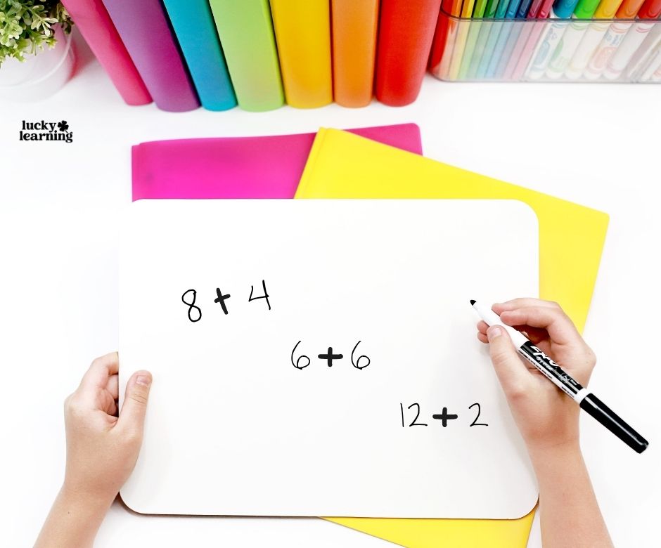 A person writes simple addition problems (8+4, 6+6, 12+2) on a whiteboard with colorful folders and markers in the background.
