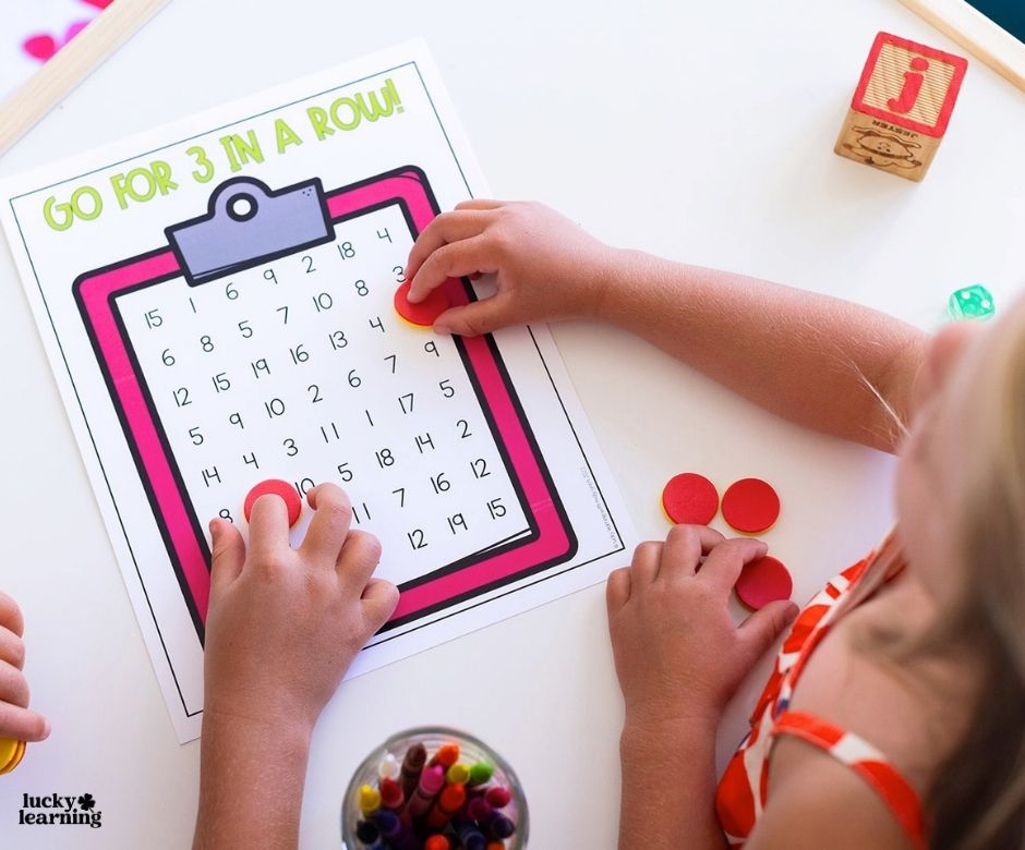 Two children play a math game called “Go For 3 in a Row!” using red counters on a numbered board with a clipboard graphic. A jar of colored pencils and wooden blocks are also on the table.