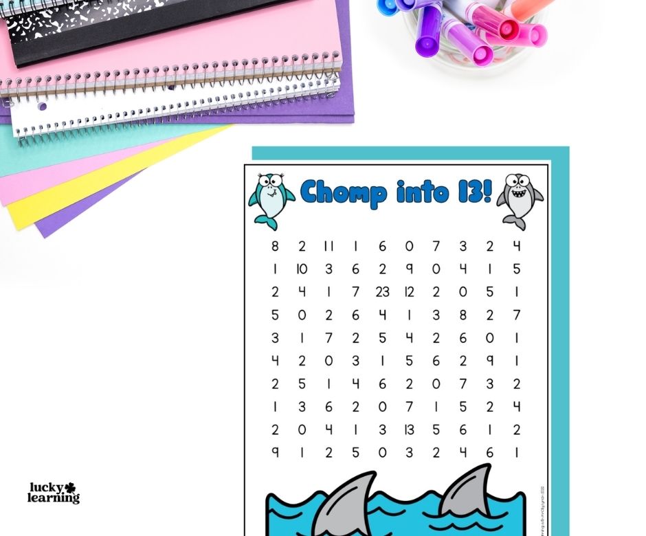 A children’s math worksheet titled “Chomp into 13!” features numbers, shark fins in water, and dolphins. Colored pens and spiral notebooks are nearby.