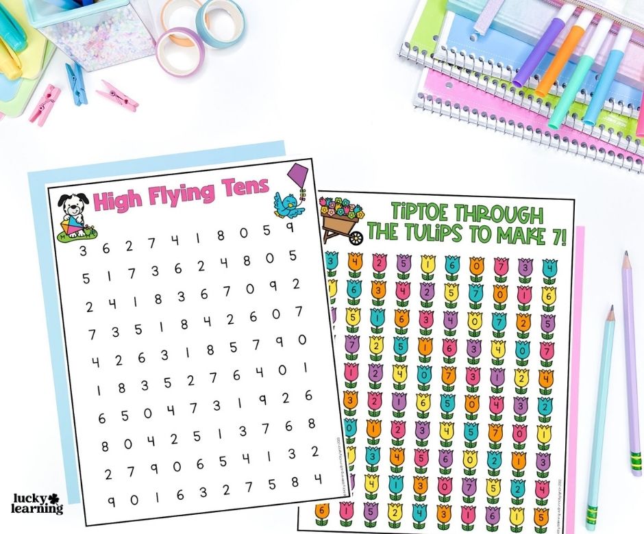 Two colorful math activity worksheets on a white desk, surrounded by pencils, pens, washi tape, and notebooks. The worksheets feature numbers and tulip illustrations.